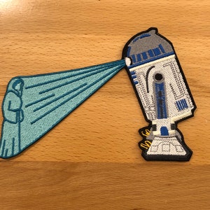 May include: An embroidered patch featuring a blue and white robot with a blue beam of light shooting out of its head. The robot is standing on a wooden surface.