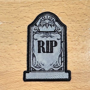 May include: Embroidered patch in the shape of a tombstone, featuring the letters "RIP" in bold, black lettering. The tombstone is gray with ornate detailing and a black border, perfect for Halloween or gothic themes.