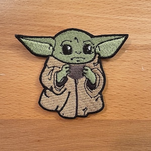 Puede incluir: Parche bordado de un personaje alienígena verde con ojos grandes, que lleva una túnica marrón. El personaje está de pie con los brazos cruzados.