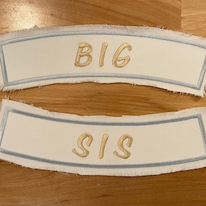 May include: Two white fabric patches with a blue border. The top patch says "BIG" in gold lettering. The bottom patch says "SIS" in gold lettering.