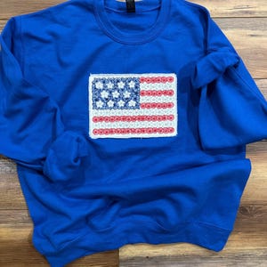 May include: A royal blue sweatshirt with an embroidered American flag design. The flag's stars are white, and the stripes are red. The sweatshirt has a crew neck and long sleeves, perfect for casual wear. The design is patriotic.