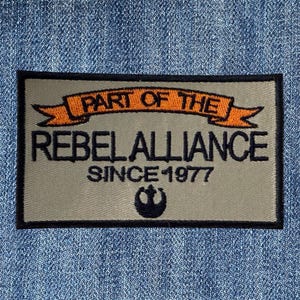 Może przedstawiać: Prostokątna naszywka z napisem "PART OF THE REBEL ALLIANCE SINCE 1977" w czarnym kolorze na beżowym tle. Tekst jest obramowany pomarańczowym banerem i czarną obwódką. Na dole znajduje się symbol Rebel Alliance.