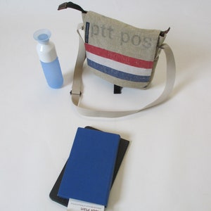 May include: A beige canvas messenger bag with a red, white and blue stripe across the front. The bag has a black strap and the text "ptt pos" printed on the front. A blue water bottle and a blue notebook with a black cover are also in the image.