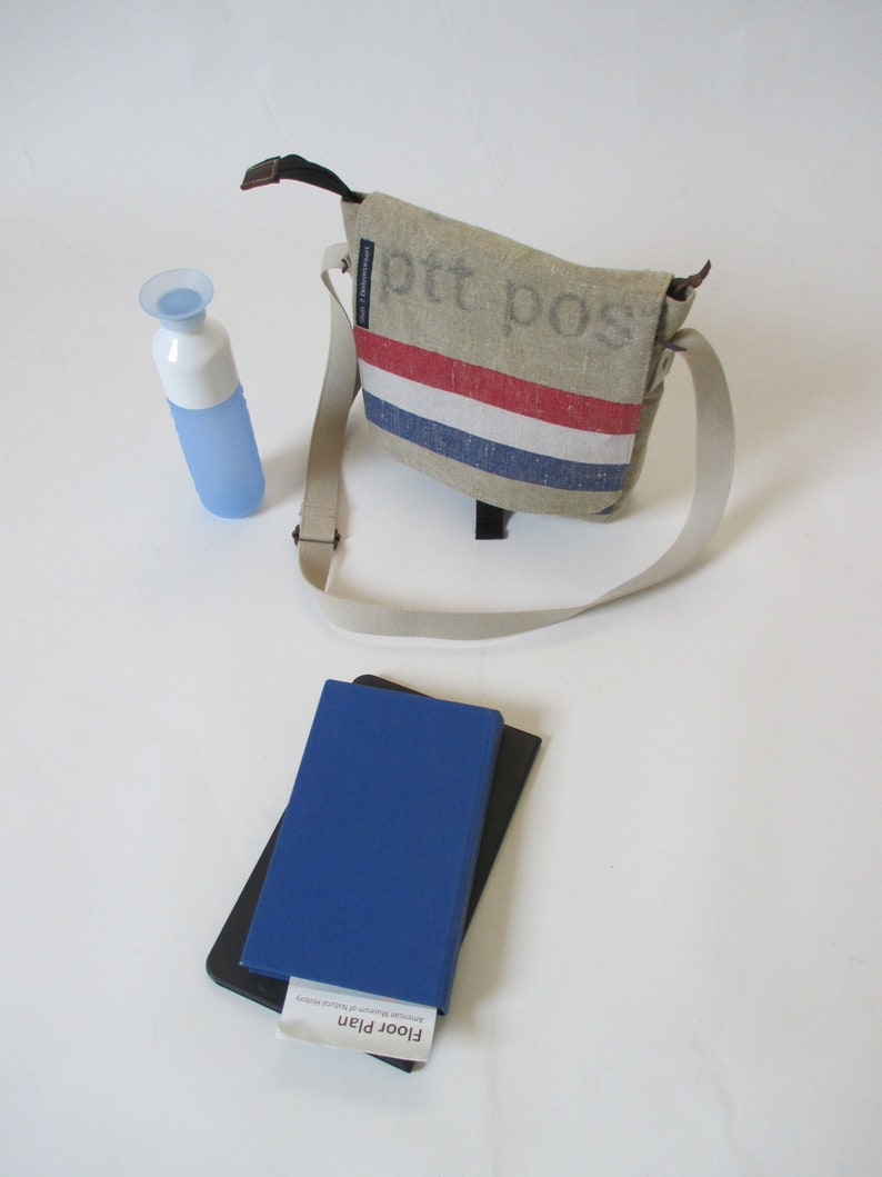 May include: A beige canvas messenger bag with a red, white and blue stripe across the front. The bag has a black strap and the text "ptt pos" printed on the front. A blue water bottle and a blue notebook with a black cover are also in the image.