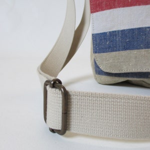 May include: A close-up of a beige canvas shoulder bag with a red, white and blue striped design. The bag has a brown metal buckle on the strap.