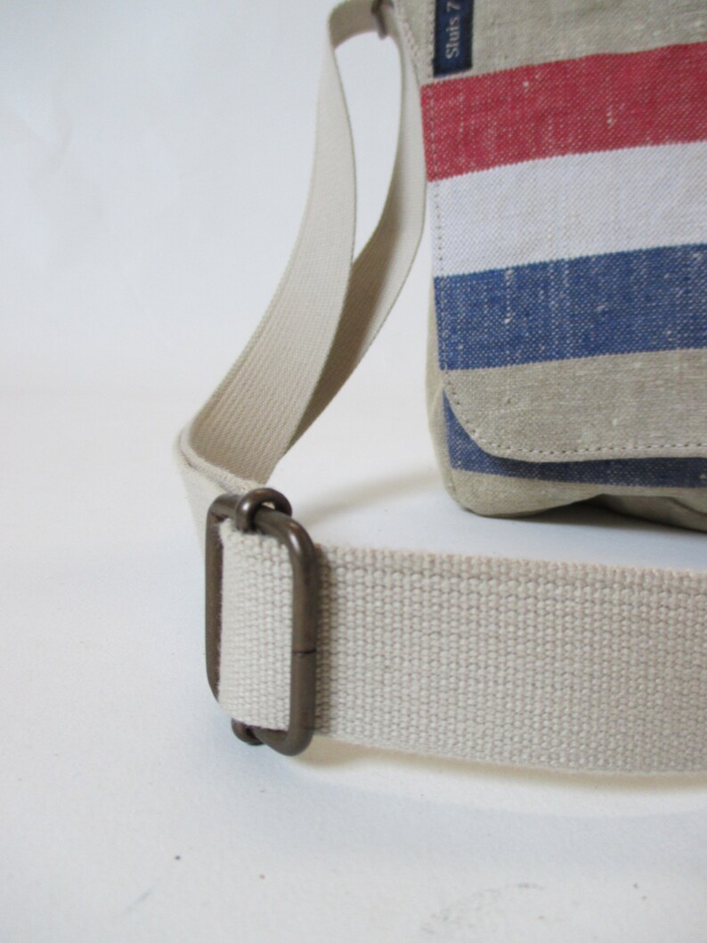 May include: A close-up of a beige canvas shoulder bag with a red, white and blue striped design. The bag has a brown metal buckle on the strap.