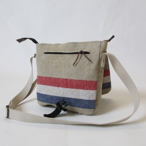 May include: A beige canvas messenger bag with a red, white and blue striped design. The bag has a zipped pocket on the front and an adjustable strap.