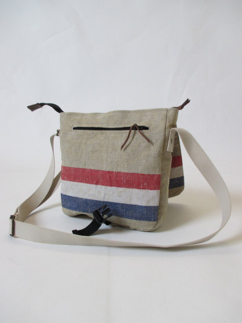 May include: A beige canvas messenger bag with a red, white and blue striped design. The bag has a zipped pocket on the front and an adjustable strap.