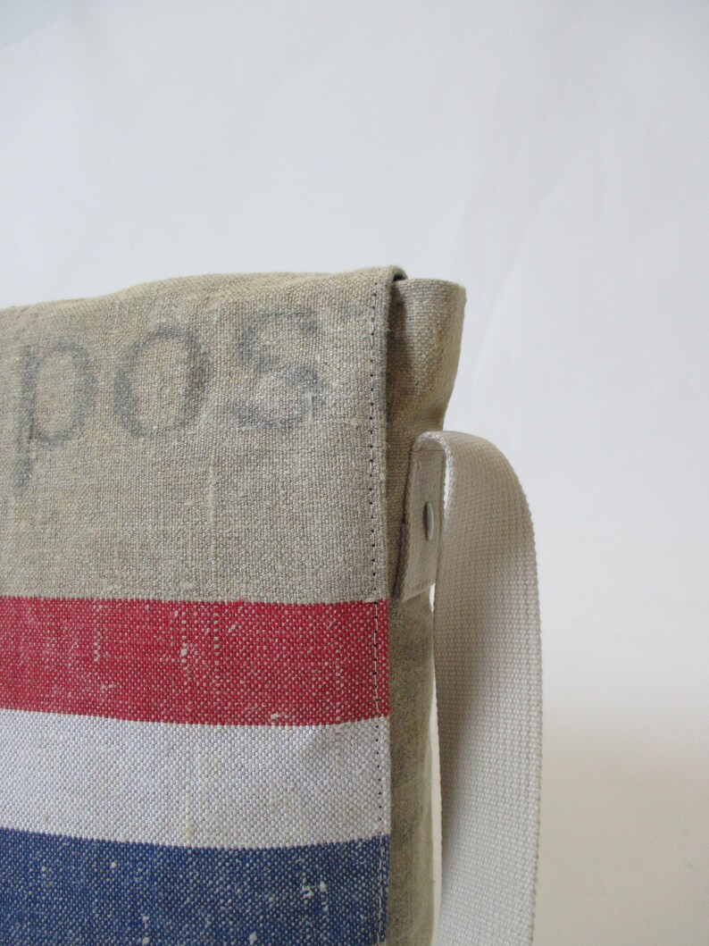 May include: A beige canvas messenger bag with a red, white and blue striped pattern. The bag has a long, adjustable strap and the word "POS" is printed on the front.