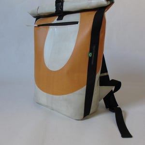 May include: A roll-top backpack with a unique design. The bag features a large orange and white pattern on the front, with black accents and straps. A zipper pocket is visible on the front. The bag is made of a durable material.