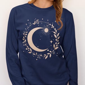 May include: A navy blue long-sleeve shirt featuring a central graphic of a large crescent moon and a small sun encircled by a wreath of leaves and small stars. The design is printed in a light beige color on the front of the garment.