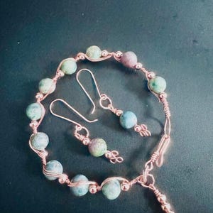 May include: A jewelry set consisting of a bracelet and a pair of drop earrings crafted from copper wire and round, multi-colored stone beads in shades of green, blue, and purple. The bracelet features a hook clasp and a small dangling charm.