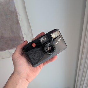 May include: Black Leica Mini Zoom camera with a textured grip and a silver lens. The camera has a built-in flash and the Leica logo in red. The lens reads "VARIO-ELMAR 35-70". The camera is held in a hand.