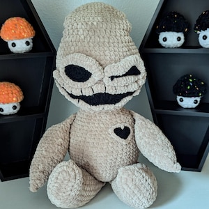 May include: A crocheted plush toy resembling a character with a stitched-on smile and a black heart. The toy is beige with black eyes and a black stitched-on mouth. The toy is sitting on a white surface.