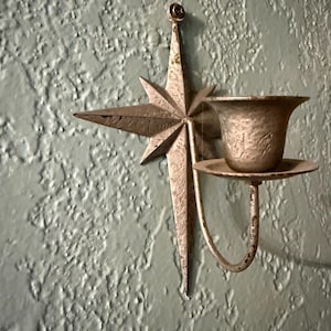 May include: A metallic wall-mounted candle holder featuring a star-shaped backplate with a weathered finish. A curved arm extends from the center of the star to support a small, flared cup designed to hold a single candle securely in place.