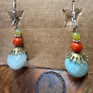 May include: A pair of dangle earrings with a light blue, rounded stone base, topped with a brass-colored filigree, a red bead, and a green bead. The earrings have silver-colored lever-back closures.
