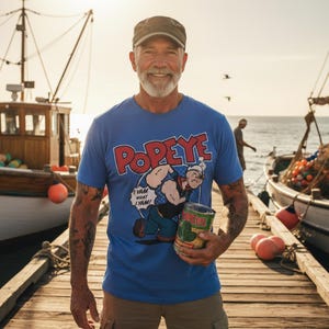May include: A blue t-shirt featuring the Popeye cartoon character with the text "Popeye" and "I yam what I yam!" A man is holding a can of spinach. The background includes boats and a pier.