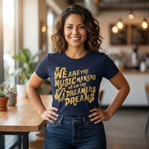 May include: A navy blue t-shirt with the text "We are the music makers, we are the dreamers of dreams" in gold lettering. The shirt is paired with blue jeans. The person is smiling and standing in a cafe.