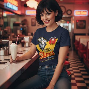 May include: A navy blue t-shirt with a vintage "Jack Rabbit Slim's" diner graphic, showing a rabbit in a suit and a milkshake. The shirt is worn with blue jeans. A milkshake with a red and white straw sits on the counter.