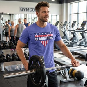 以下が含まれることがあります： ジムで、青いTシャツを着た人がいます。「AMERICAN GLADIATORS」の文字が白と赤でプリントされています。Tシャツのデザインには星と赤と白の三角形が含まれています。その人はバーベルを持っています。他のジムの利用者と設備が見えます。