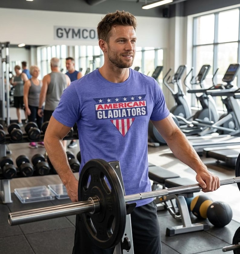 以下が含まれることがあります： ジムで、青いTシャツを着た人がいます。「AMERICAN GLADIATORS」の文字が白と赤でプリントされています。Tシャツのデザインには星と赤と白の三角形が含まれています。その人はバーベルを持っています。他のジムの利用者と設備が見えます。