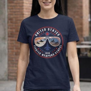 Puede incluir: Camiseta azul marino con un diseño circular. El diseño dice "UNITED STATES FIGHTER WEAPONS SCHOOL 1986" y presenta aviones de combate reflejados en gafas de sol de aviador. El diseño es en rojo, blanco y azul.