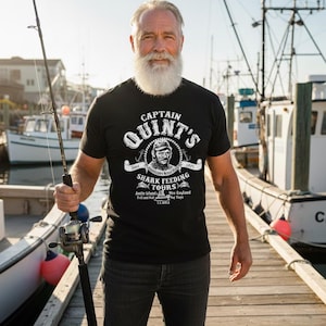 Puede incluir: Una camiseta negra con el texto "CAPTAIN QUINT'S SHARK FEEDING TOURS" y un gráfico de la cara de un hombre. La persona de la imagen sostiene una caña de pescar en un muelle de madera con barcos al fondo.
