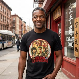 May include: Black t-shirt featuring a movie poster design from "The Last Dragon" in a circular frame. The poster has a vintage aesthetic with various characters and the movie title in red. The person is wearing blue jeans.