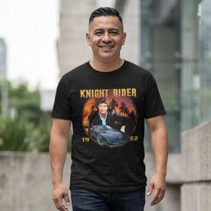 May include: Black t-shirt featuring a graphic from the television show Knight Rider. The design includes the show's title, a picture of the car, and the years 1982. The person in the image is wearing the shirt.