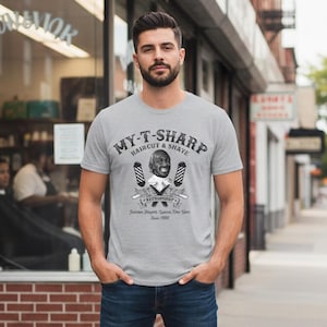 May include: A light grey t-shirt with the text "MY-T-SHARP Haircut & Shave" and a barber shop graphic. The graphic includes a portrait, barber poles, and the text "BEERLISHIP". The person is wearing blue jeans.