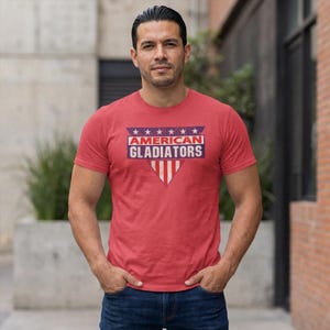 A male model wearing a heather red t-shirt with the American Gladiator logo on his chest. This is a tribute design from the 80s tv show called "American Gladiator"