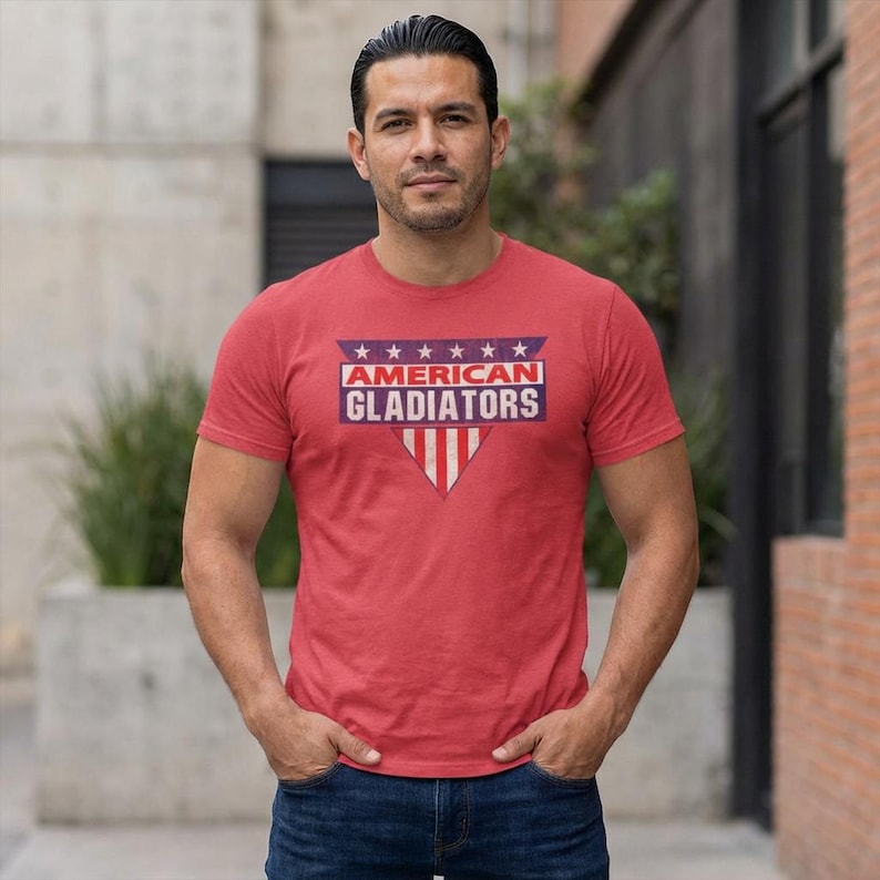 A male model wearing a heather red t-shirt with the American Gladiator logo on his chest. This is a tribute design from the 80s tv show called "American Gladiator"