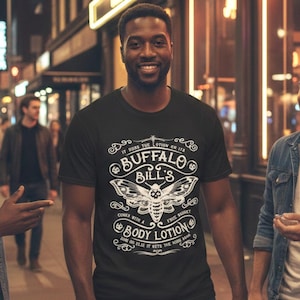 May include: Black t-shirt with a white graphic design featuring the text "Buffalo Bill's Body Lotion" and a moth illustration. The shirt also includes the phrases "It rubs the lotion on its skin" and "Comes with a free basket."