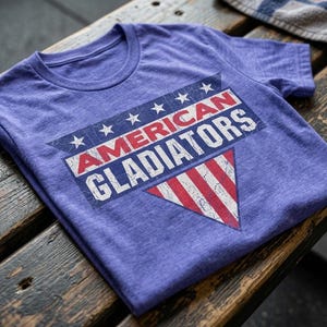 以下が含まれることがあります： 「AMERICAN GLADIATORS」の文字がプリントされた紫色のTシャツ。古びた愛国的なデザインで、赤と白のストライプと白い星が特徴です。ヴィンテージ感を醸し出しています。木製の表面に折りたたまれています。