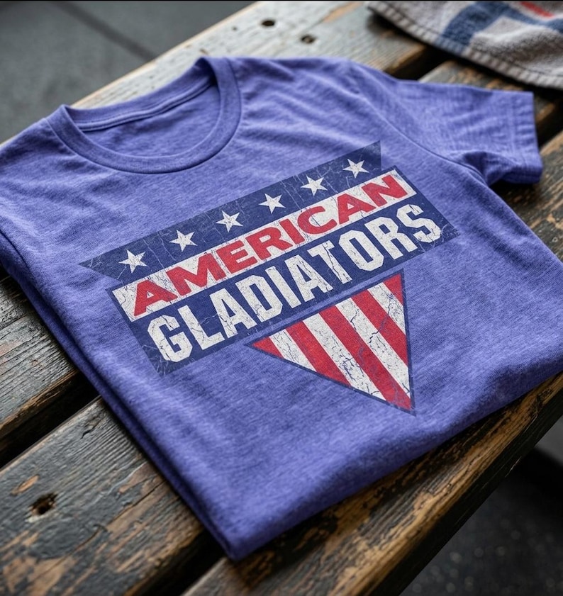 以下が含まれることがあります： 「AMERICAN GLADIATORS」の文字がプリントされた紫色のTシャツ。古びた愛国的なデザインで、赤と白のストライプと白い星が特徴です。ヴィンテージ感を醸し出しています。木製の表面に折りたたまれています。