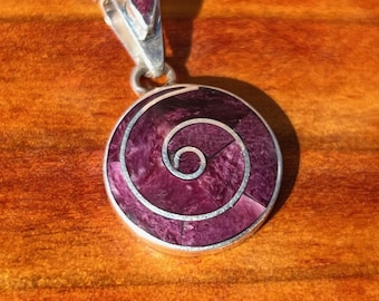 May include: A circular pendant featuring a deep purple stone inlay arranged in a spiral pattern. The pendant is framed in silver and includes a silver bail at the top, which is also accented with a small, matching purple stone inlay detail.