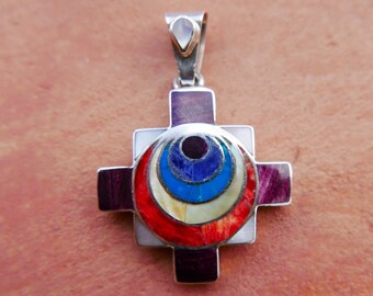 May include: A silver pendant in the shape of a cross with a colorful circular design in the center. The design features concentric circles of red, white, blue, and purple.