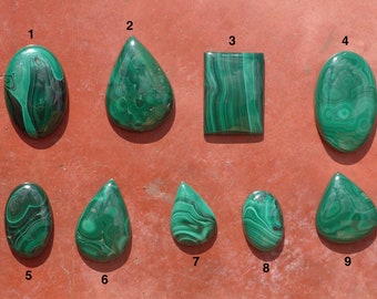 Puede incluir: Nueve cabujones de malaquita verde en varias formas y tamaños, incluyendo ovalado, gota y rectangular. Los cabujones están dispuestos en dos filas sobre una superficie marrón rojiza.