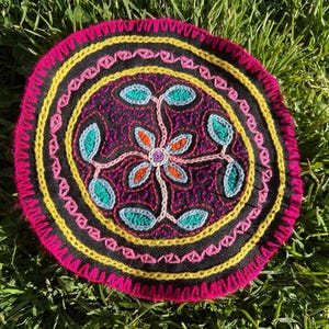 Può includere: Tessuto circolare ricamato con sfondo nero e motivo floreale centrale. Il design presenta accenti turchese, arancione e viola, circondati da bordi gialli e rosa. Il tessuto è posto su erba verde.