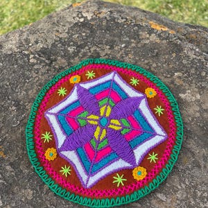 Op de afbeelding: Een kleurrijk, cirkelvormig, geborduurd ontwerp met een geometrisch patroon in paars, wit, groen, roze en blauw. Het ontwerp is omgeven door een bruine rand met gele bloemen en een groen-roze-gele gehaakte rand.