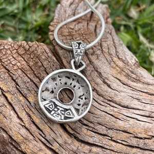 May include: A silver-toned pendant necklace with a circular design and intricate cut-out patterns. The pendant hangs from a thin, silver chain. The pendant has a central hole and a detailed design. The necklace is displayed on a piece of wood.