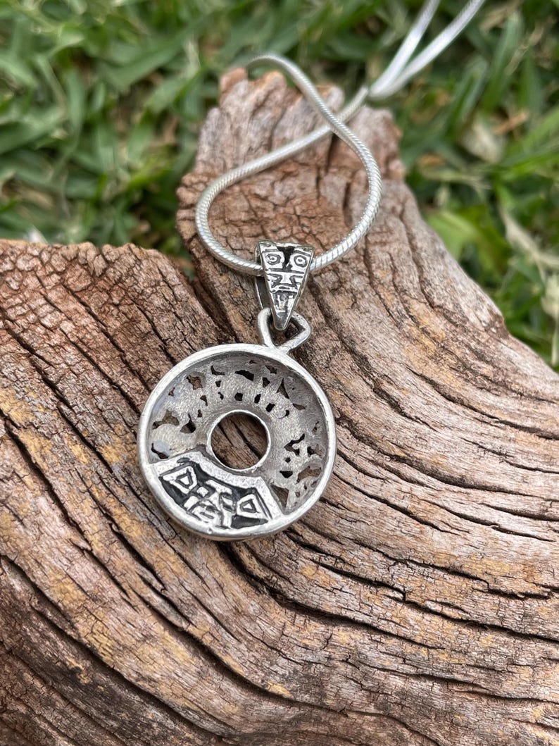 May include: A silver-toned pendant necklace with a circular design and intricate cut-out patterns. The pendant hangs from a thin, silver chain. The pendant has a central hole and a detailed design. The necklace is displayed on a piece of wood.