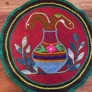 Op de afbeelding: Een kleurrijk rond wandkleed met borduurwerk met een vaas waaruit twee slangen komen. De vaas is versierd met een bloem en strepen. De achtergrond is bruin met een groene, gele en zwarte rand.