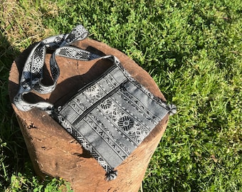 May include: A gray and black patterned crossbody bag with a matching strap. The bag features a zippered main compartment and a front pocket. The strap and bag have a geometric design. The bag is resting on a wooden surface.