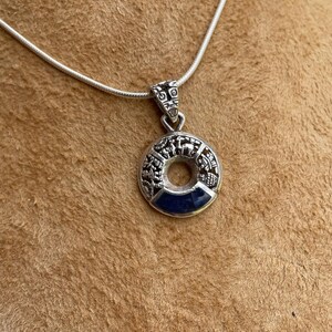 May include: A silver pendant necklace featuring a circular pendant with intricate relief details. The pendant has a dark blue center and is suspended from a delicate silver chain. The design appears to be inspired by ancient architecture.