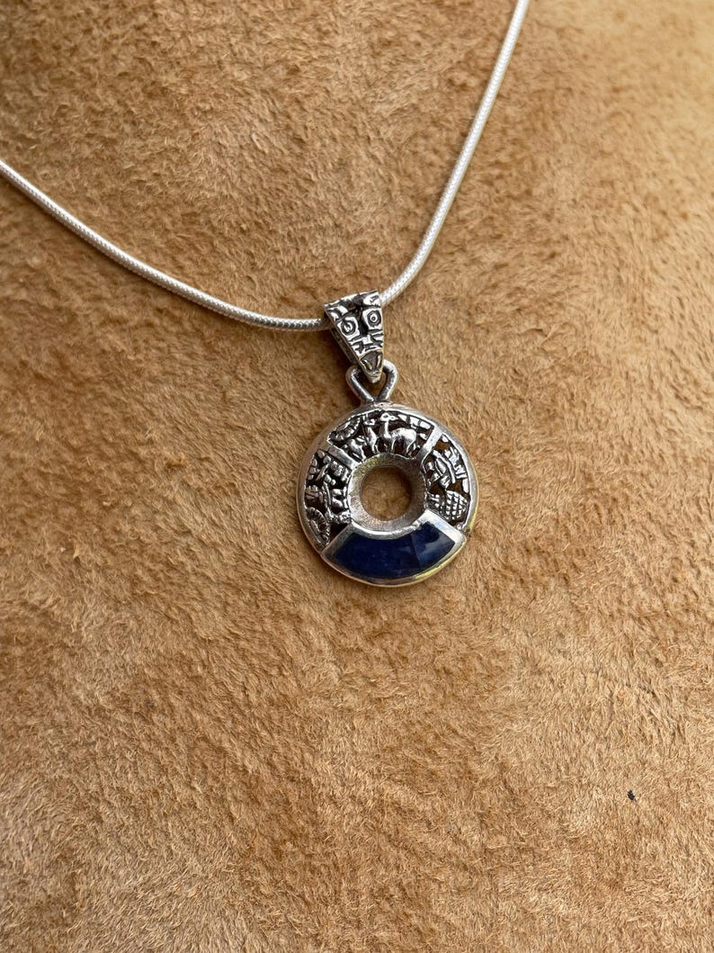 May include: A silver pendant necklace featuring a circular pendant with intricate relief details. The pendant has a dark blue center and is suspended from a delicate silver chain. The design appears to be inspired by ancient architecture.