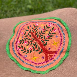 Puede incluir: Un parche de tela de color melocotón con un borde verde, que presenta un árbol bordado con flores rosas, hojas verdes y estrellas amarillas.