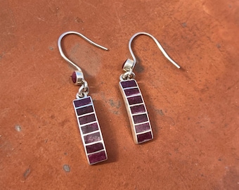 May include: A pair of silver drop earrings featuring a rectangular pendant design. Each earring consists of a silver frame inlaid with six small, deep purple rectangular stone segments. A small round purple stone sits above the main pendant.
