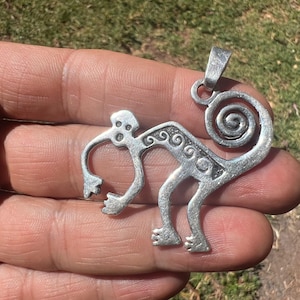 May include: A silver-toned metal pendant shaped like a stylized monkey with a curled tail. The monkey figure features etched spiral and circular patterns along its back and limbs, with a small loop at the top for attachment to a necklace chain.