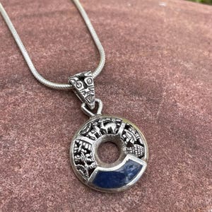 May include: A silver pendant necklace featuring a circular pendant with intricate carvings and a dark blue stone inlay. The pendant hangs from a delicate silver chain, showcasing a detailed design with a central hole.